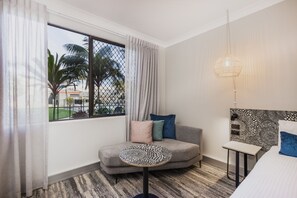 Relax in a stylish room with a comfortable sofa, a neatly made bed, and a large window offering a view of palm trees and a building.