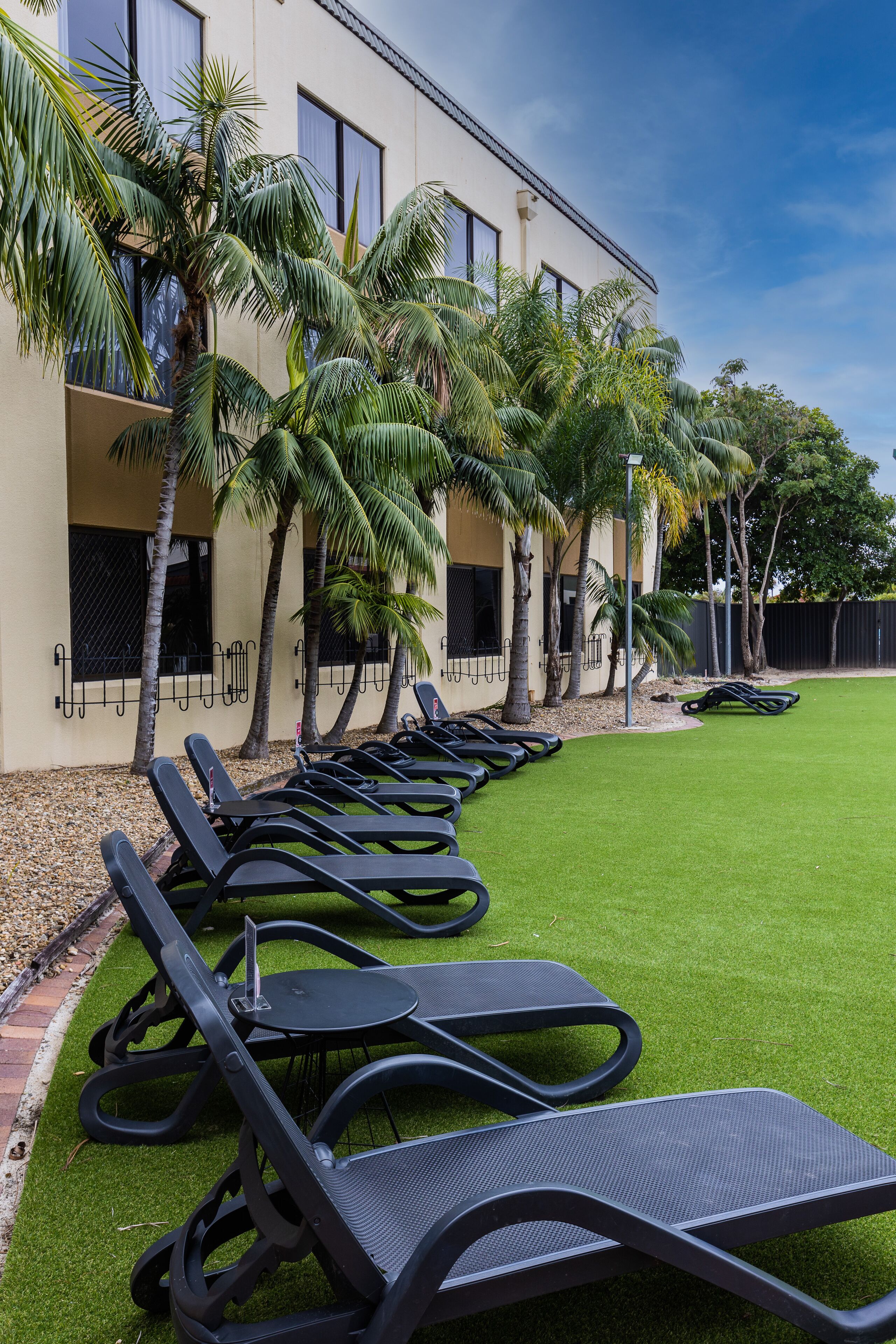 A row of black lounge chairs on a green lawn, a beige building with windows, and palm trees.