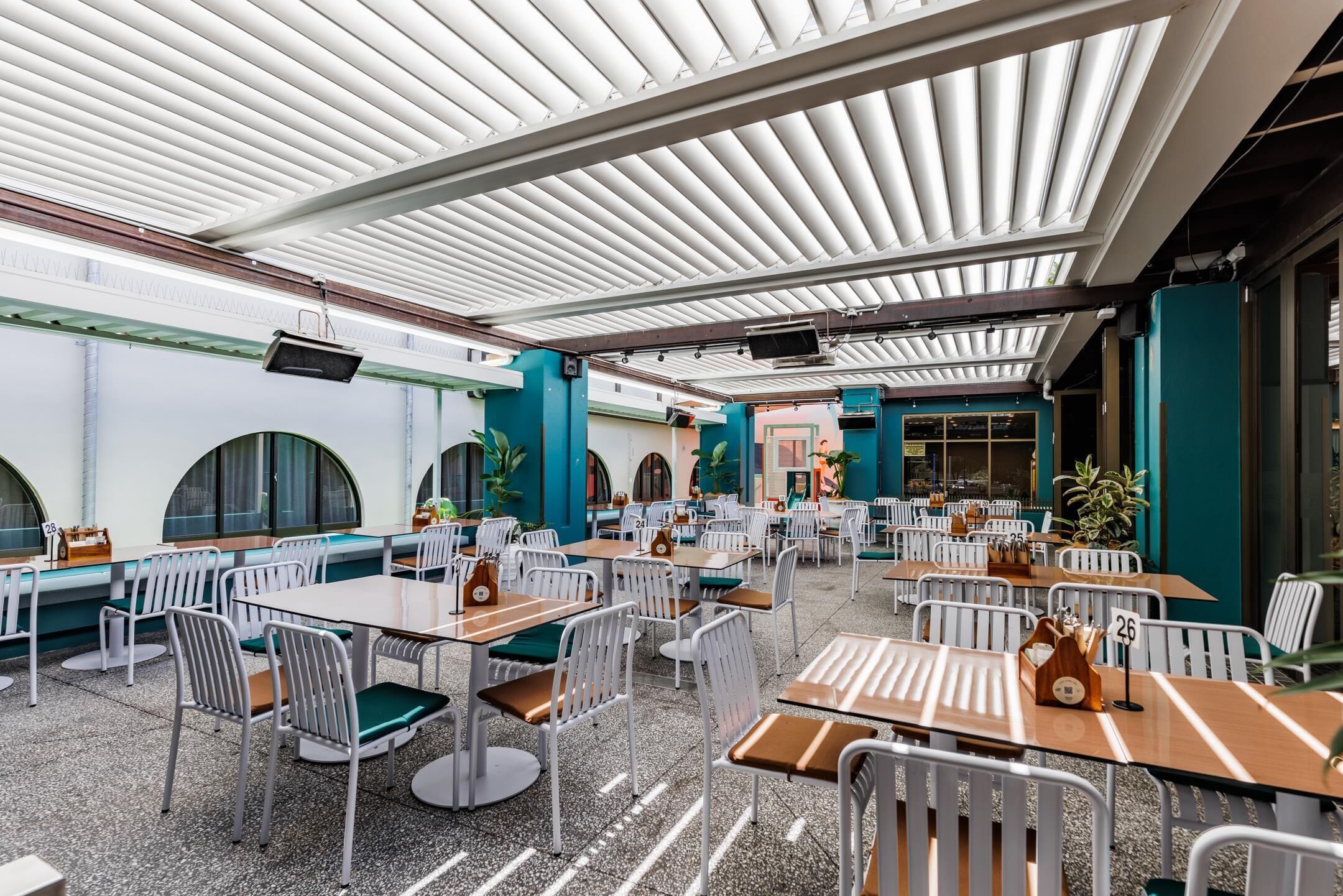 A spacious indoor dining area with white tables and chairs, a high ceiling with a patterned design, and a view of the street through large windows.