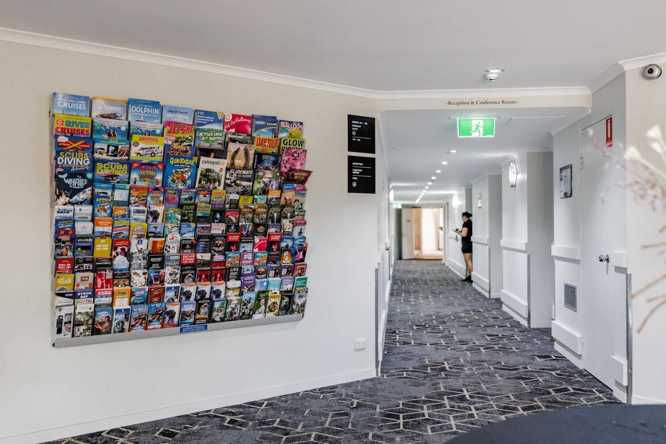 A hotel corridor with a wall displaying various travel brochures. There are reception and conference rooms visible in the background.