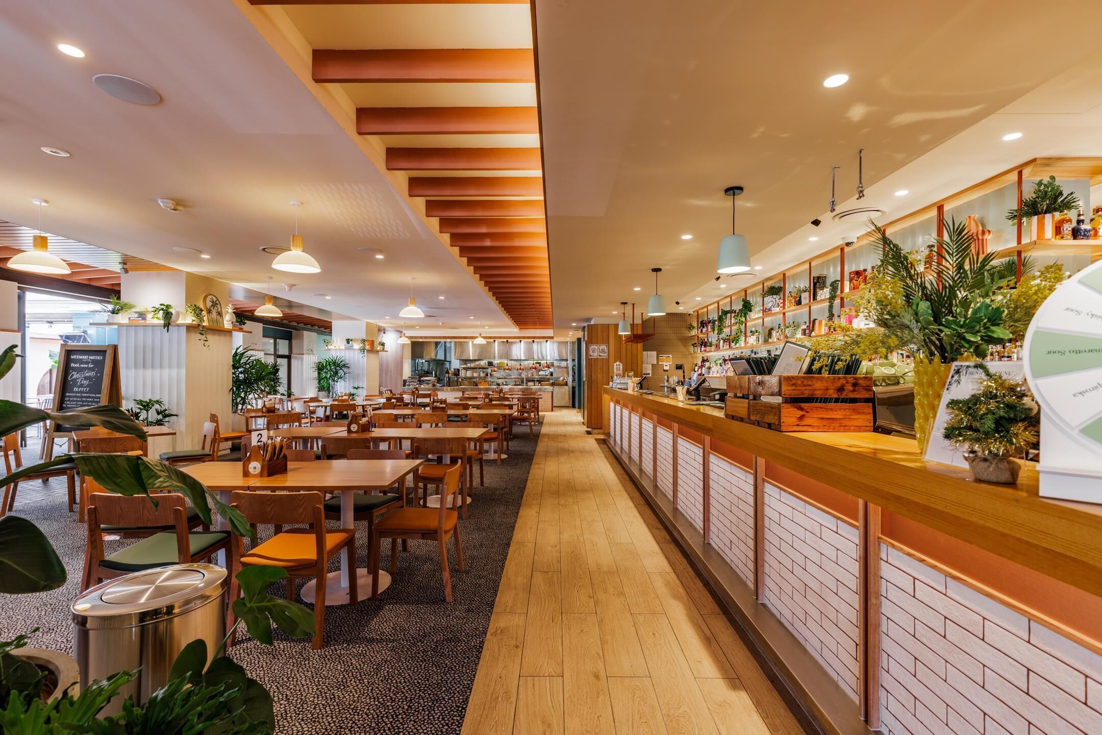 A restaurant interior with wooden tables and chairs, a counter with plants, and a kitchen in the background.