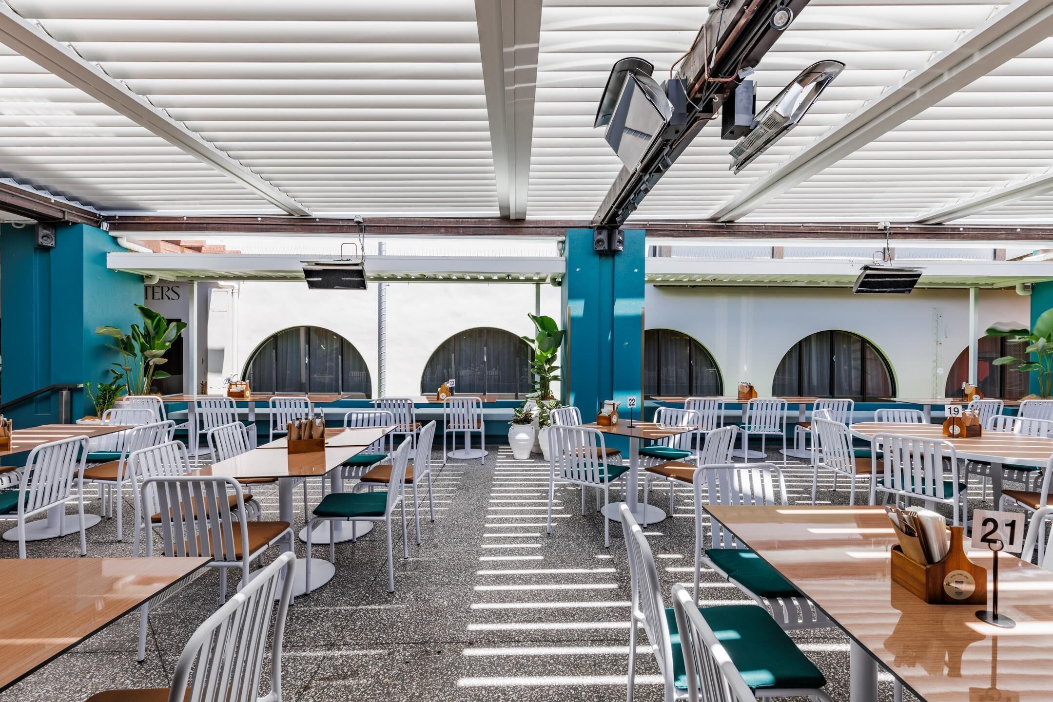 Un vaste espace extérieur aménagé pour les repas, avec des chaises et des tables en métal blanc, un sol en gravier et un plafond couvert.