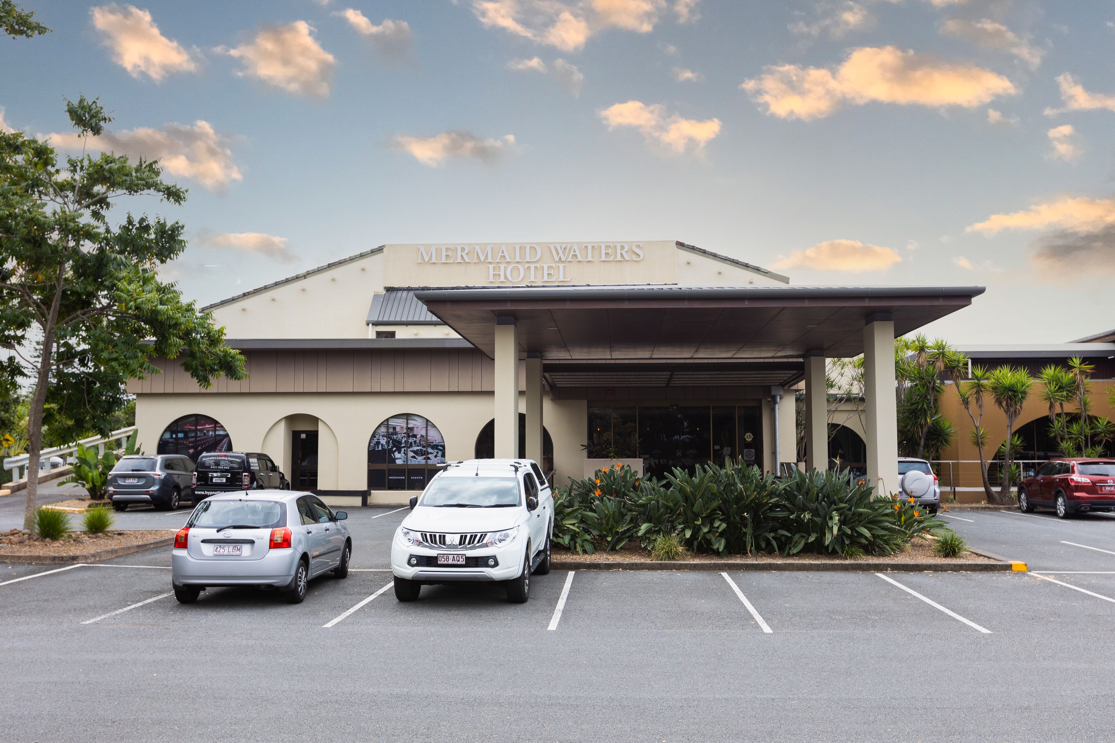 The exterior of the Mermaid Waters Hotel with a covered entrance, parking lot, and a clear sky.