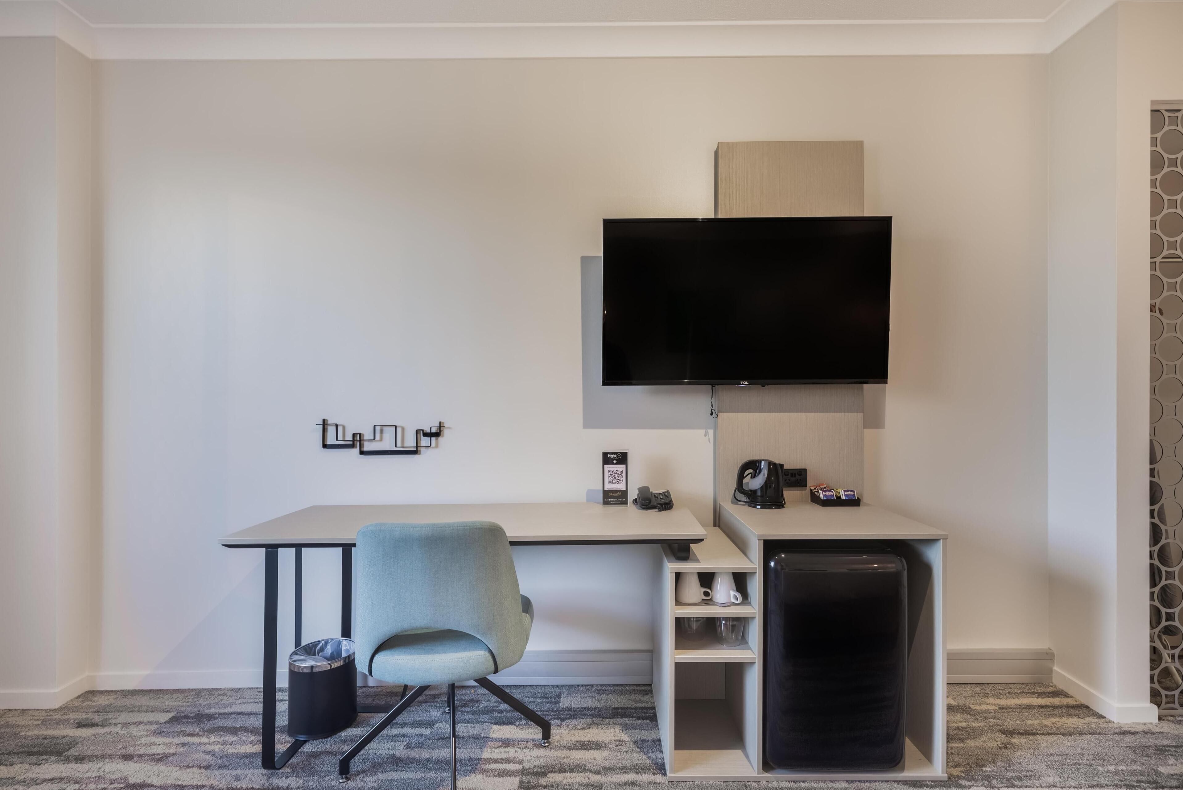 A hotel room with a desk, chair, TV, and a coffee machine.