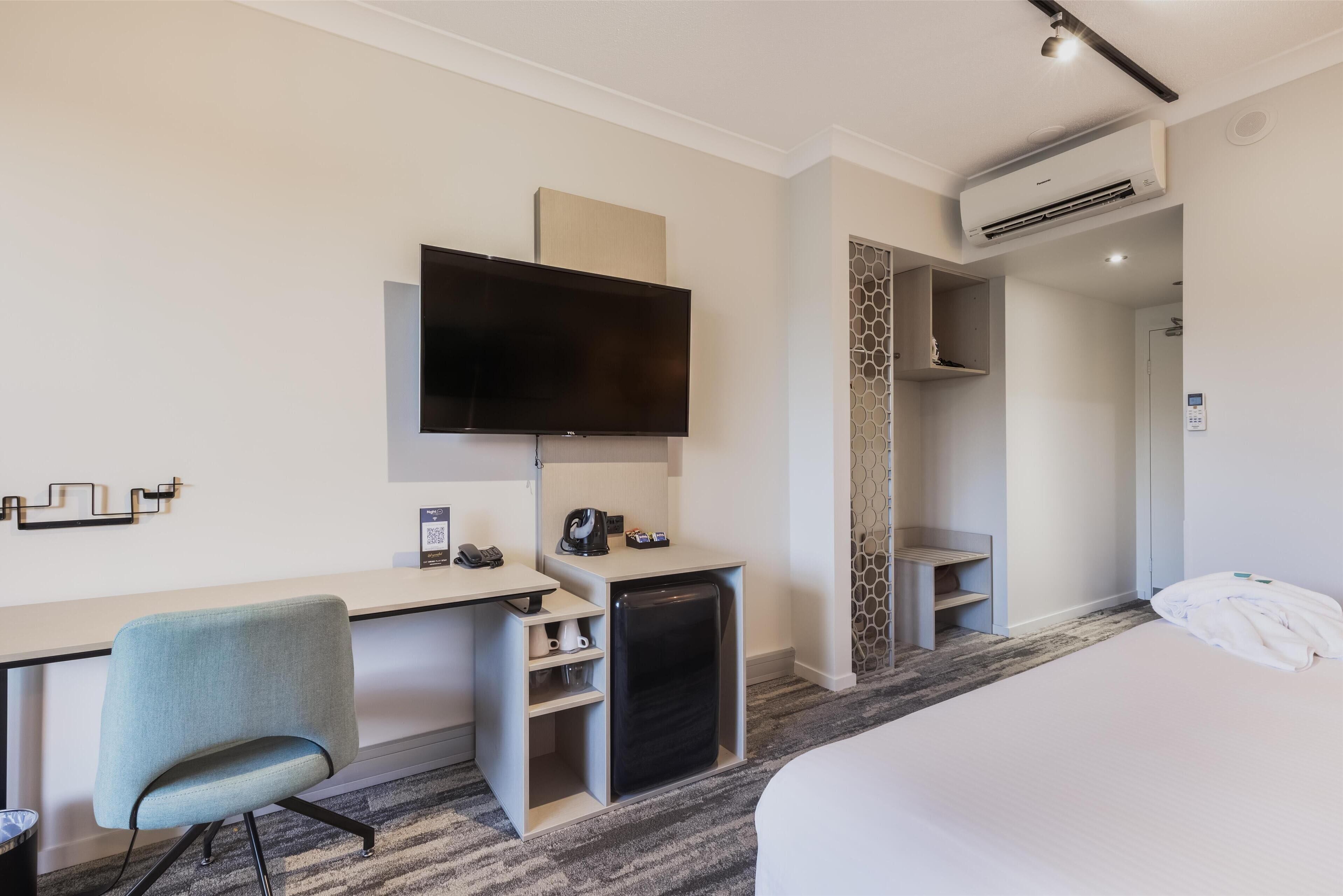 A hotel room with a bed, a desk with a chair, a television, a mini-fridge, and an air conditioning unit.
