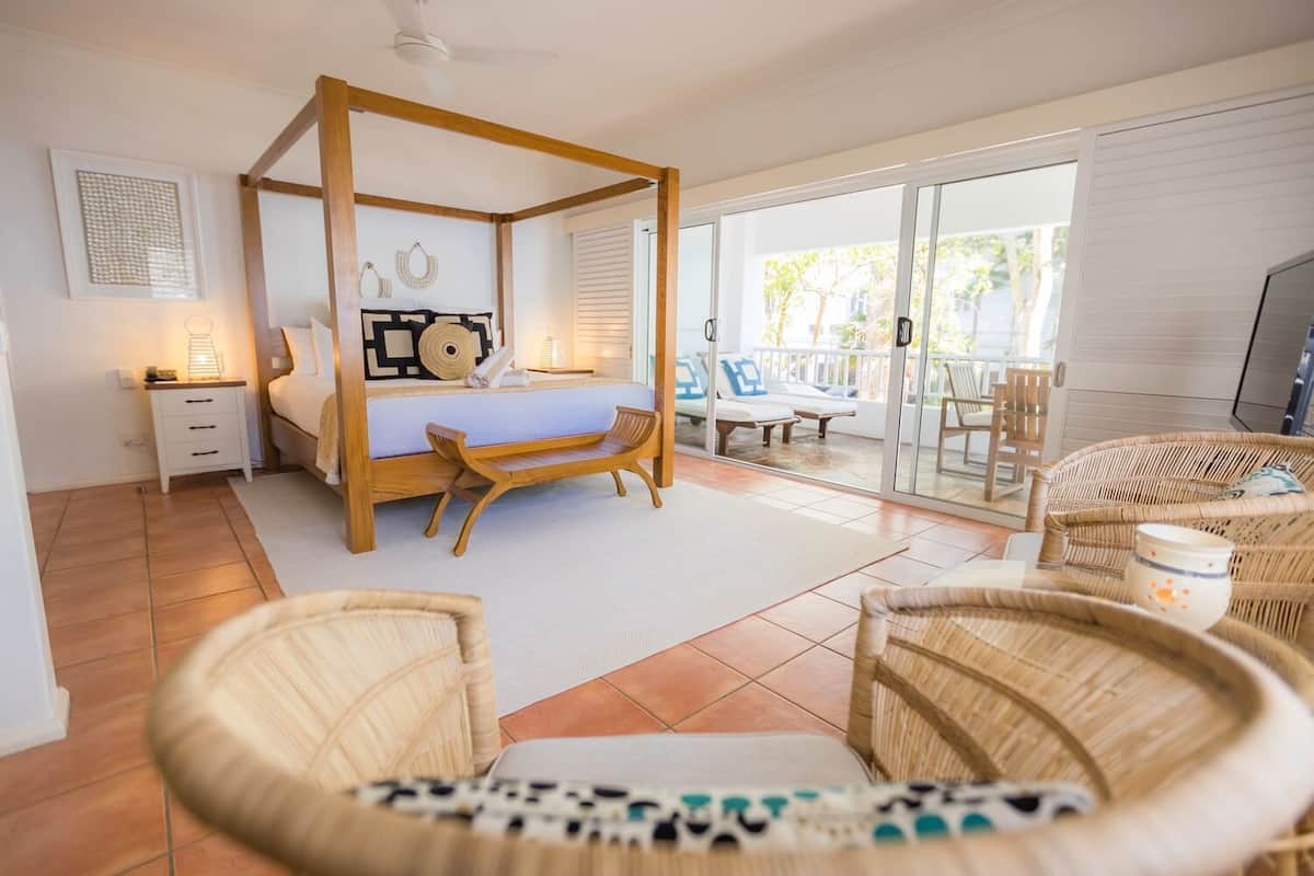 A spacious bedroom with a four-poster bed, a sitting area with wicker furniture, and a balcony with outdoor seating.