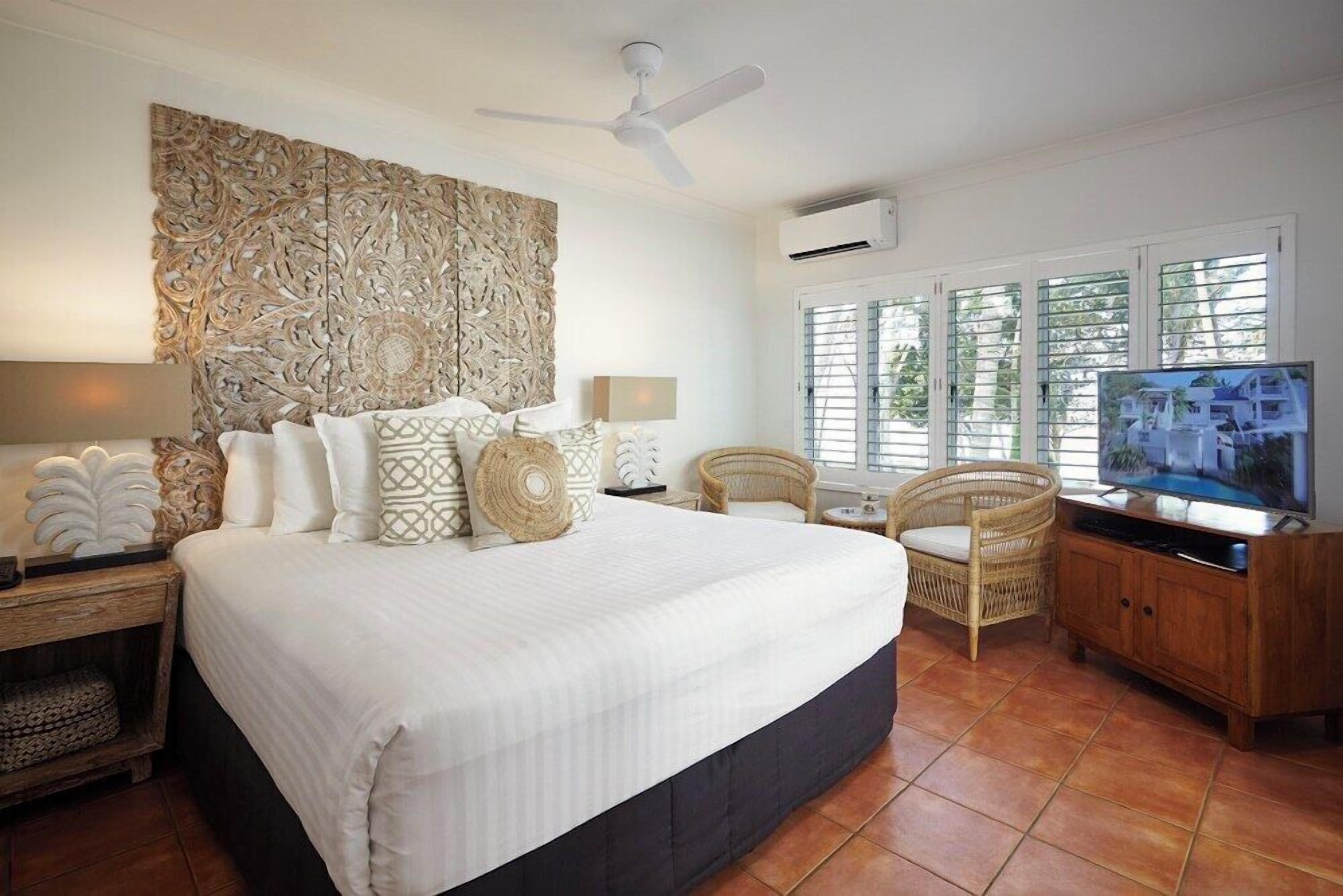 A spacious bedroom with a large bed, a ceiling fan, a TV, and a window with blinds.