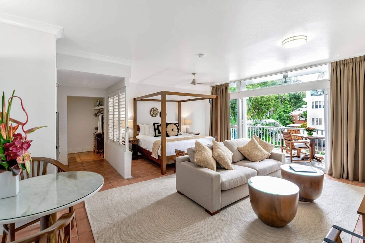 A hotel room with a balcony, a bed with a canopy, a sofa, a coffee table, and a dining area with a glass table.
