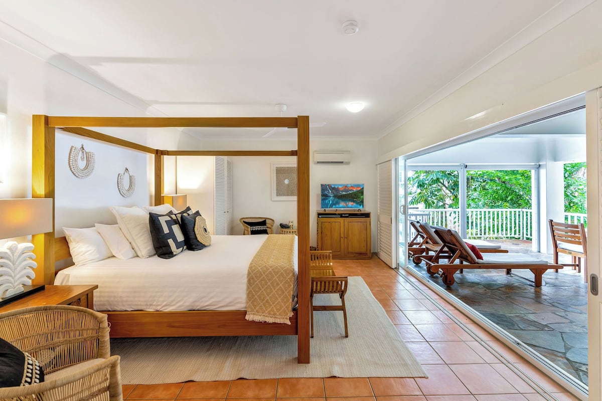 A four-poster bed with a wooden frame, a white headboard, and a beige throw blanket. There is a wicker chair and a small table with a lamp. A large window with a view of greenery is visible.