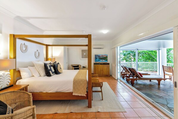 A four-poster bed with a wooden frame, a white headboard, and a beige throw blanket. There is a wicker chair and a small table with a lamp. A large window with a view of greenery is visible.