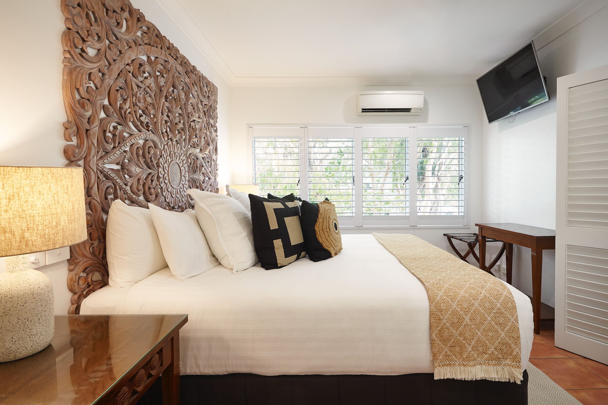 A bedroom with a large bed, a wooden headboard, a TV, an air conditioner, and a window with blinds.
