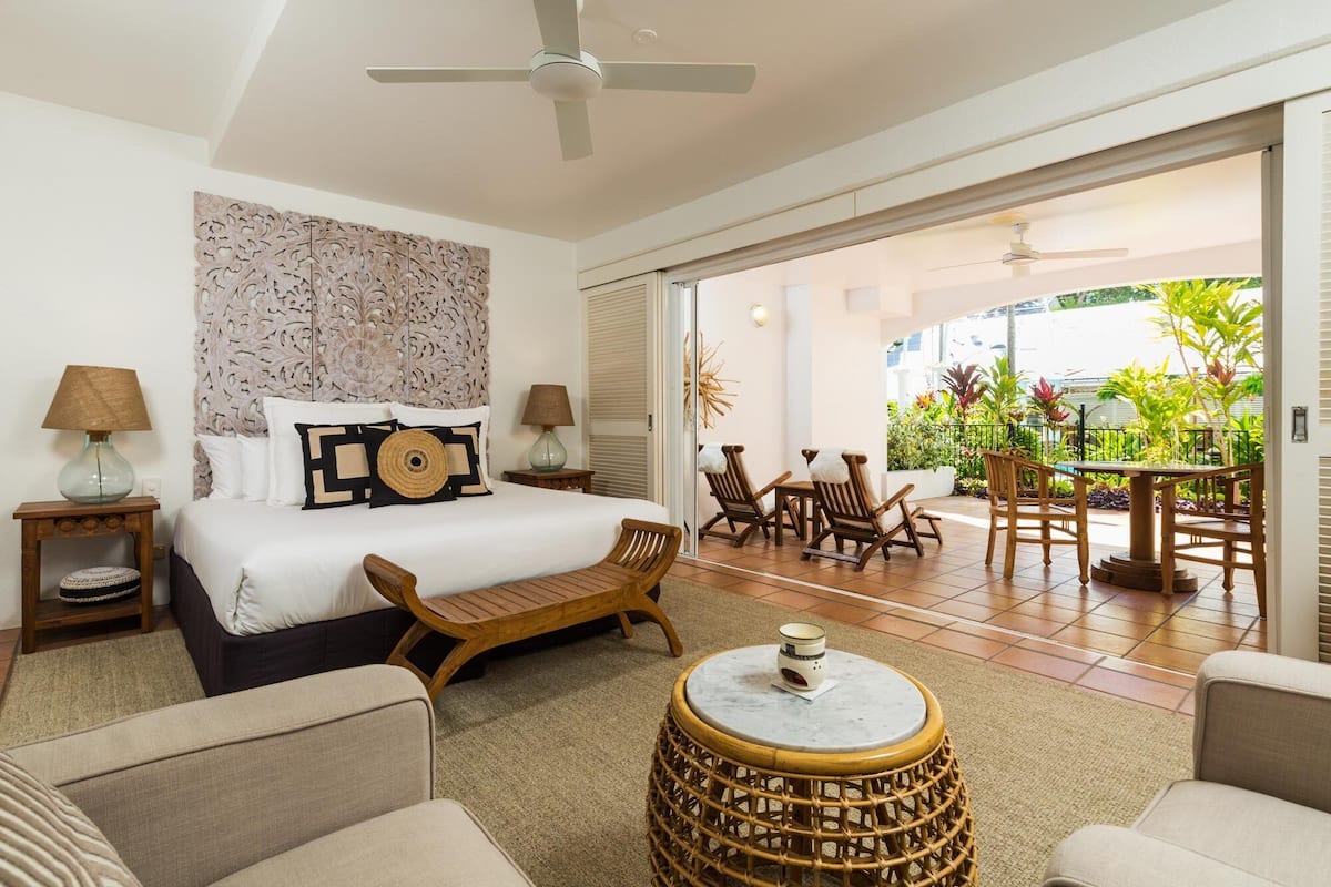 A hotel room with a bed, a small table, a chair, a round wicker stool, and a view of the outdoor patio with chairs and a table.