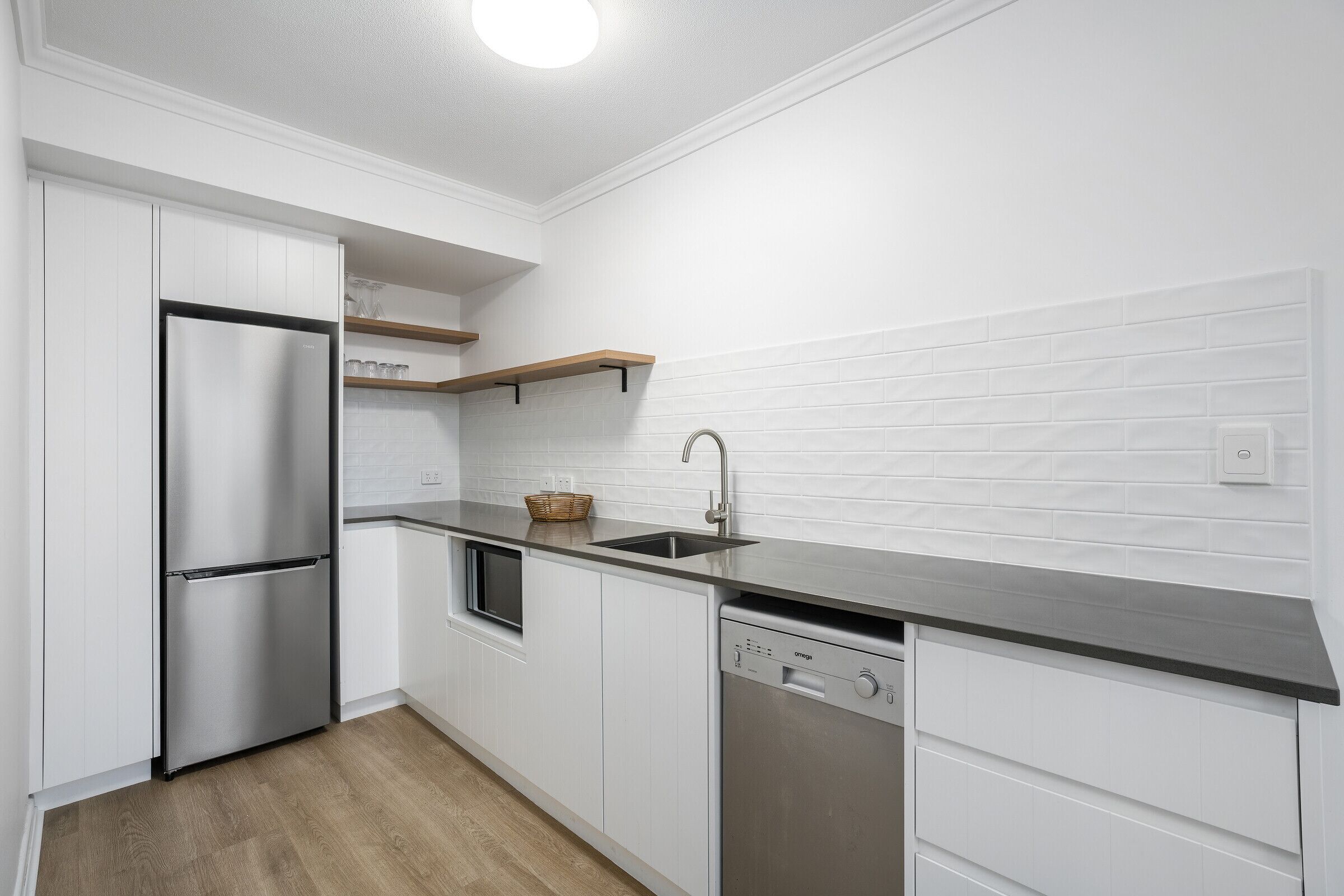 Eine moderne Küche mit weißen Schränken, einem Edelstahlkühlschrank, einem Geschirrspüler und einem hölzernen Regal über der Spüle.