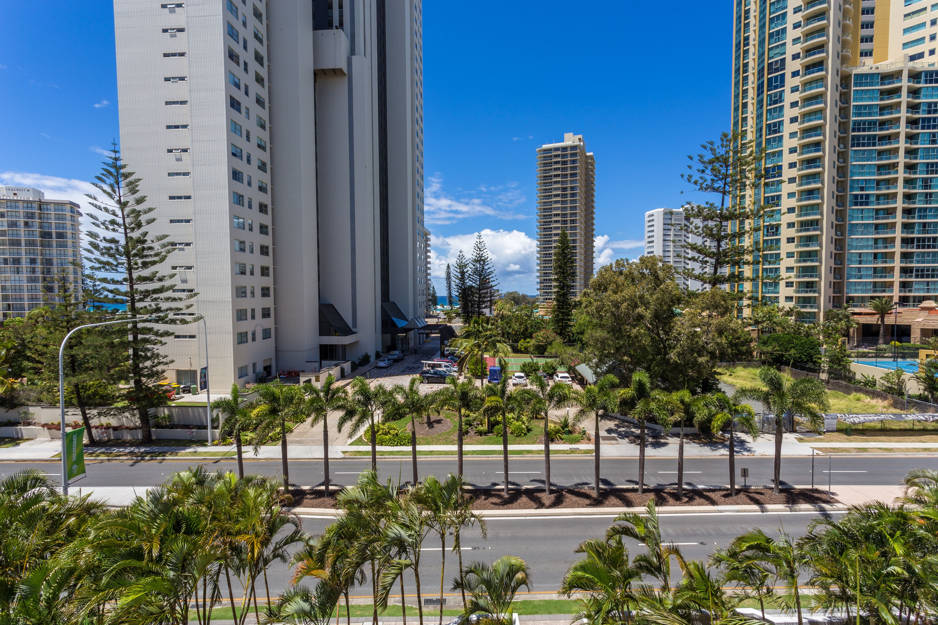 A modern urban street with tall buildings, palm trees, and a clear blue sky.