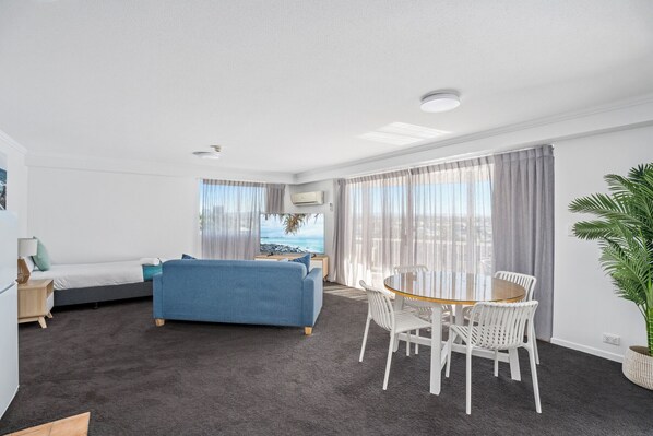 A modern hotel room with a blue sofa, a wooden dining table with white chairs, a large potted plant, and a view of the sea through the window.
