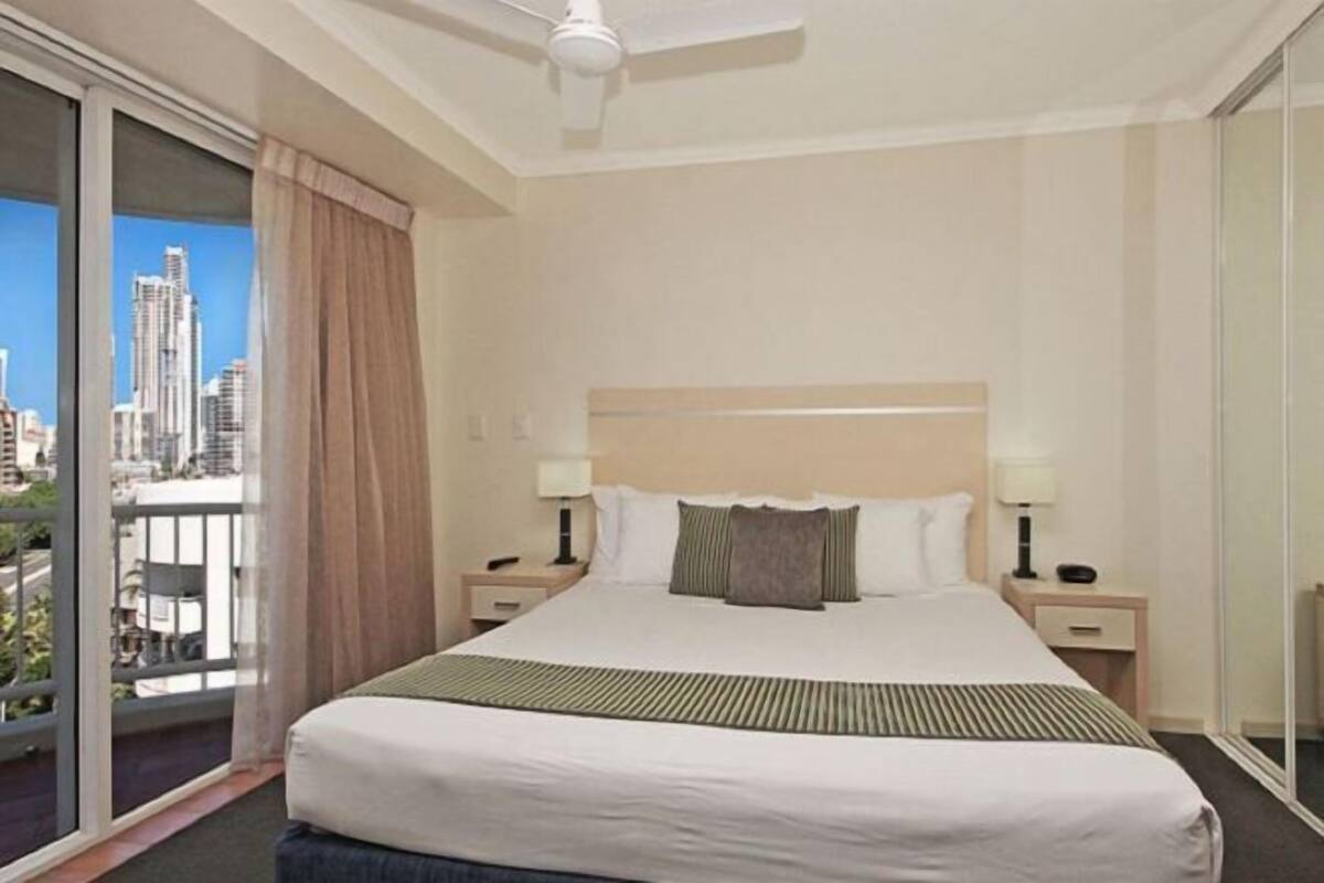 Et hotelværelse med en stor seng, en ventilator i loftet, et natbord, en lampe og udsigt til bylandskab gennem en skydedør.