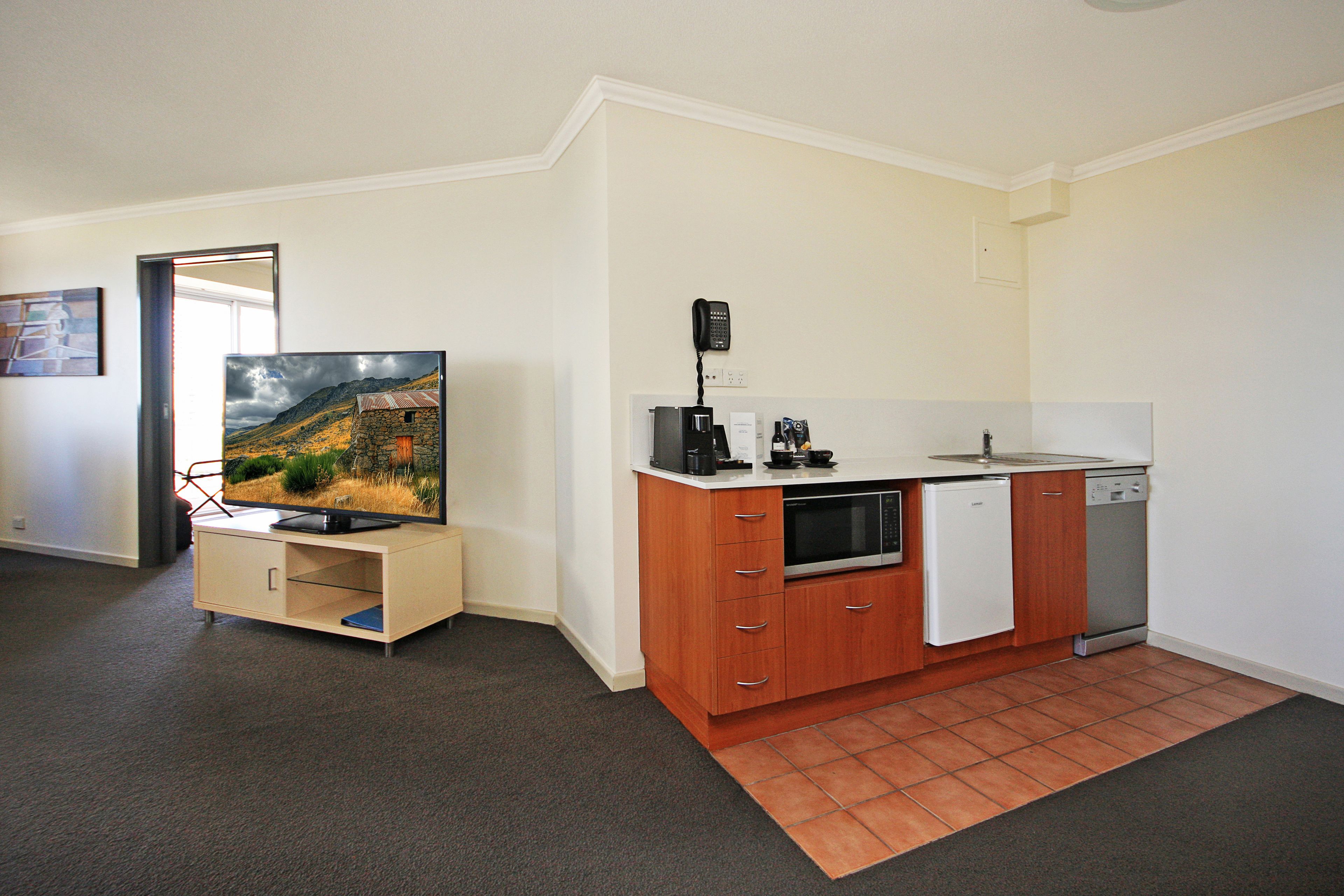 A compact kitchen with a built-in oven, microwave, and dishwasher, a flat-screen TV on a wooden cabinet, and a small countertop with a coffee maker.