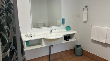 Baño con lavamanos blanco, espejo y toallero. Hay un cubo de basura y una cortina de ducha con estampado de hojas.