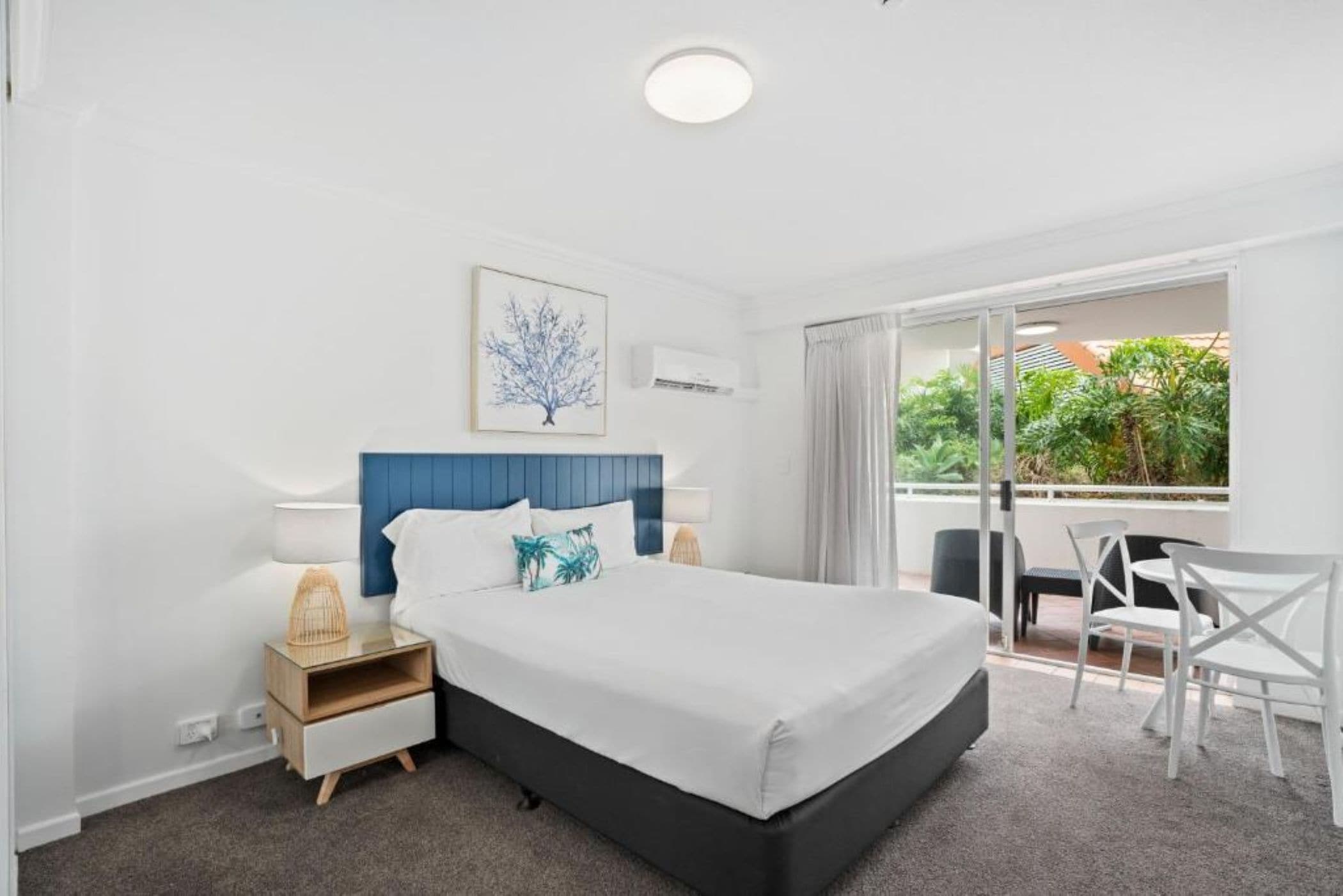A hotel room with a bed, bedside tables, a desk, and a chair. There is a window with a view of greenery outside.