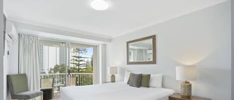 A hotel room with a large bed, a bedside table with a lamp, a chair, and a balcony with a view of trees.
