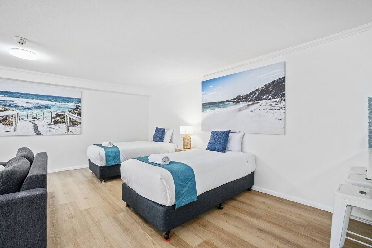 Et hotelværelse med to senge, en sofa, et lille bord og billedkunst med motiver fra kysten.