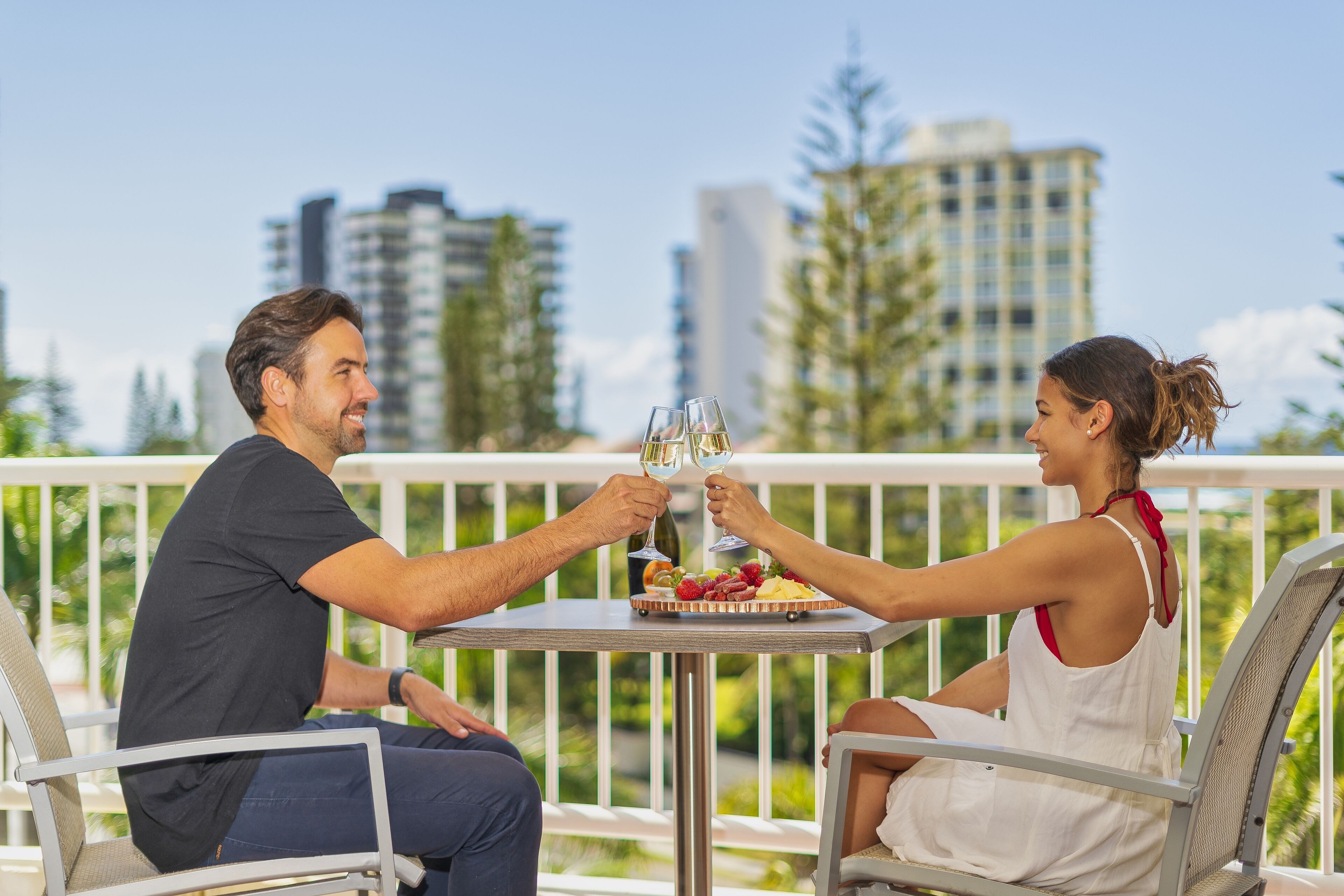 Una pareja brindando con copas de vino en una mesa con una bandeja de frutas, con vistas a un paisaje urbano con edificios altos y árboles.