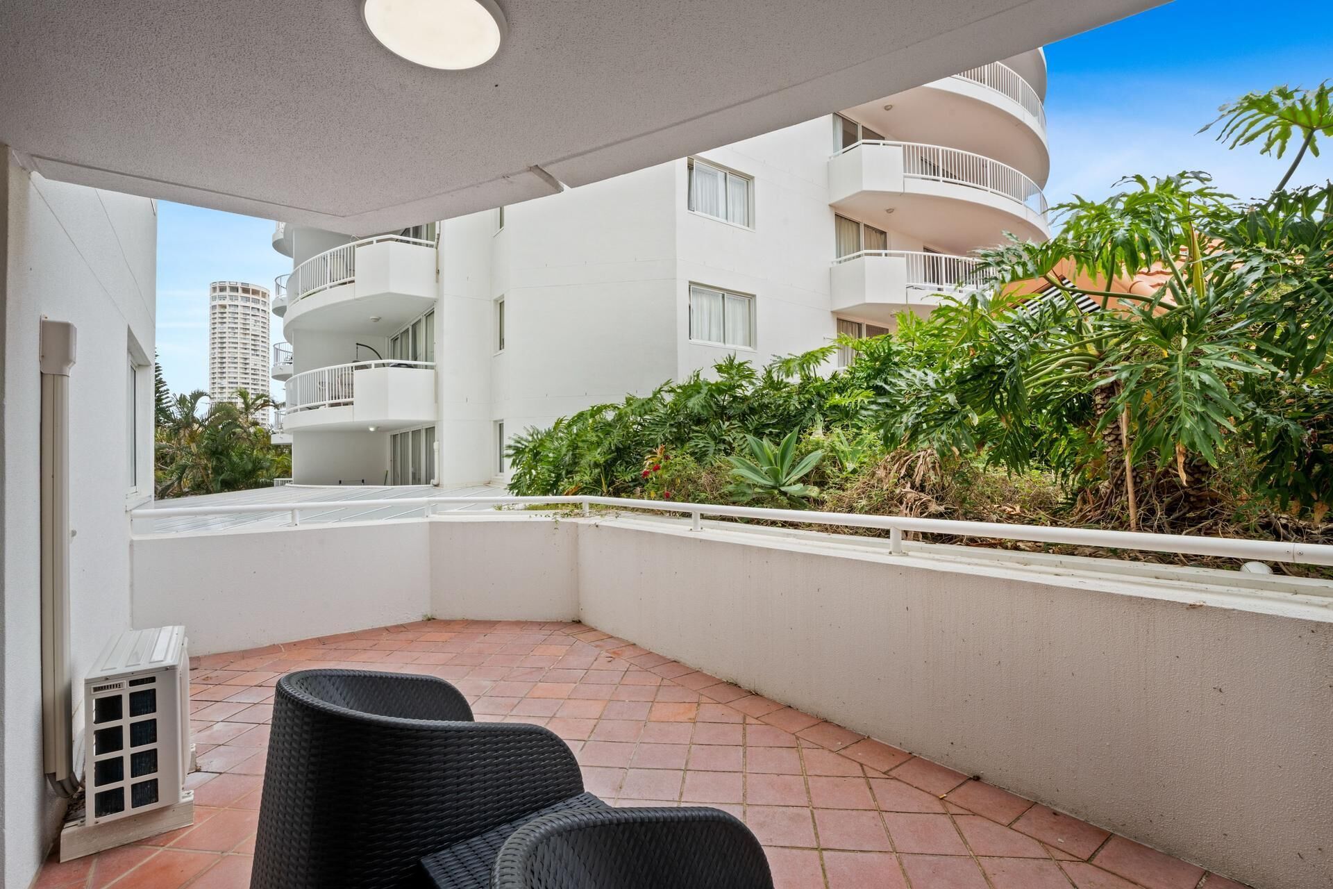 A balcony with two wicker chairs, a white air conditioning unit, and a view of a white building with multiple balconies and lush greenery.