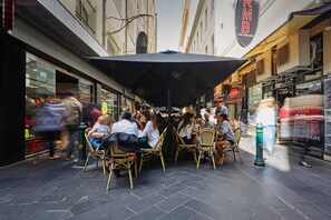 Um café ao ar livre com cadeiras de vime e um grande guarda-sol, situado numa rua estreita com lojas de ambos os lados.