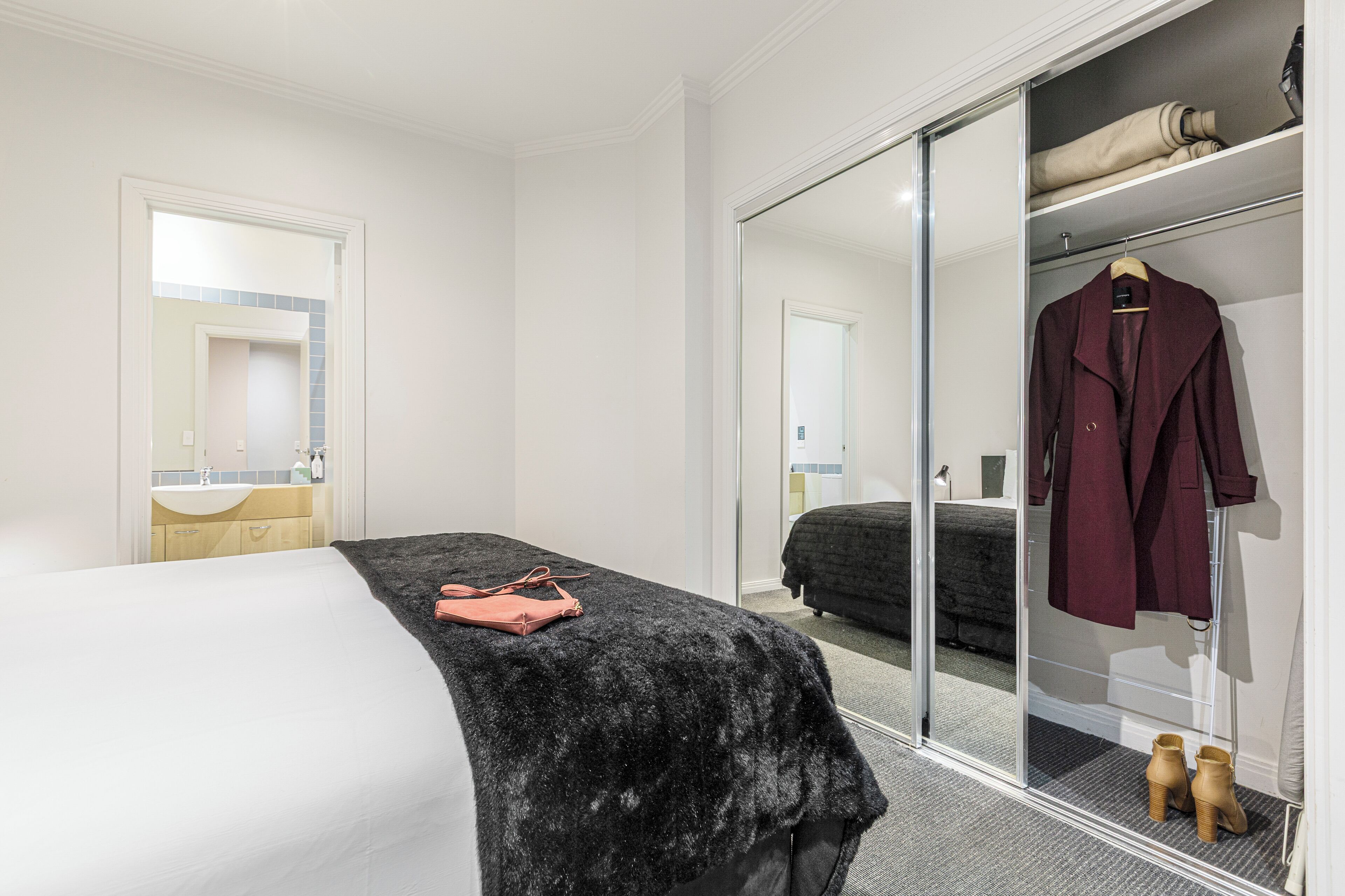 A hotel room with a bed, a black furry throw, a pink bag, a wardrobe with a hanging coat, and a mirror reflecting the room.