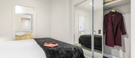 A hotel room with a bed, a black furry throw, a pink bag, a wardrobe with a hanging coat, and a mirror reflecting the room.