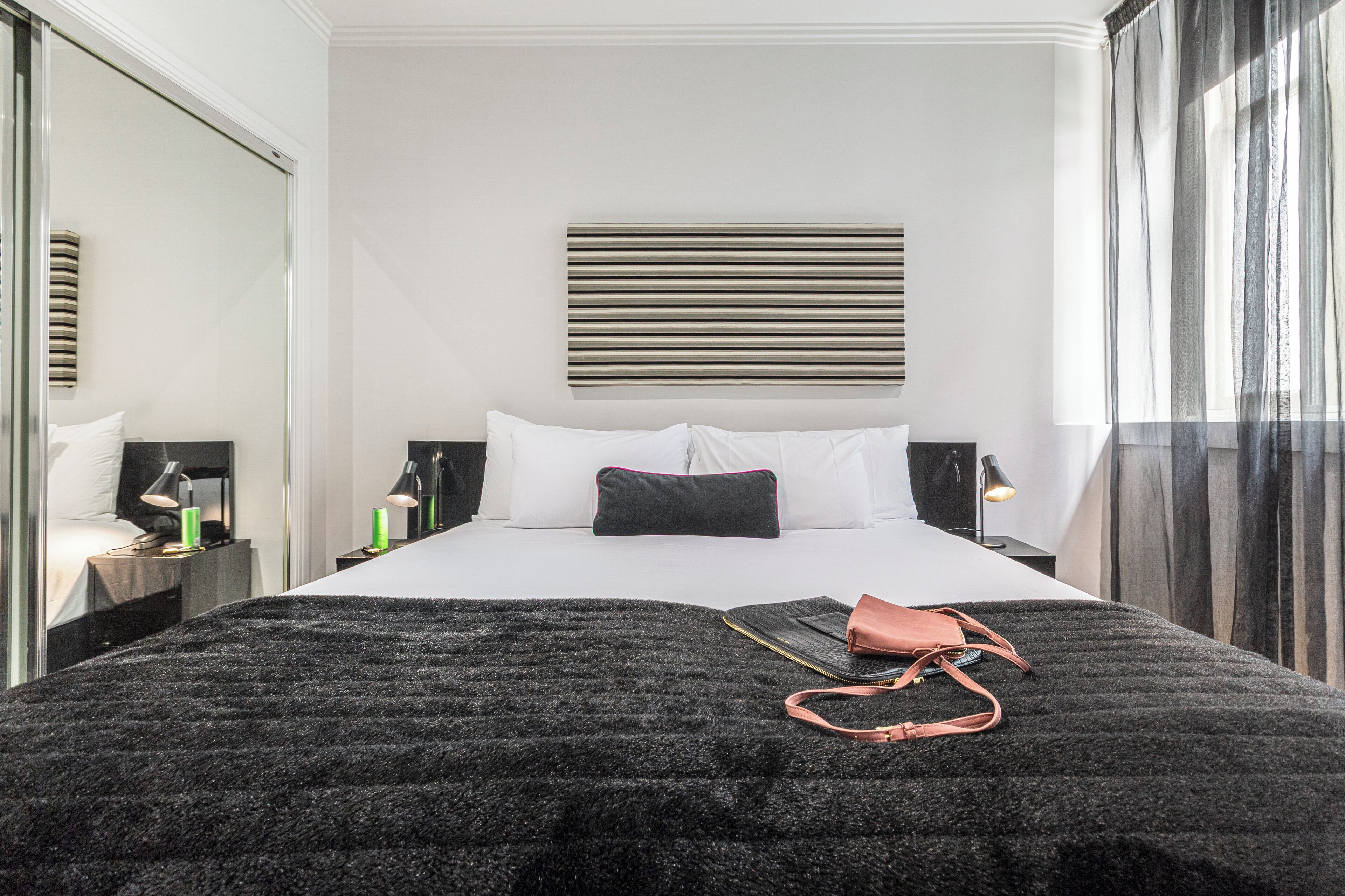 Una habitación de hotel moderna con una cama grande, una manta negra con textura, un bolso rosa y una mesita de noche con una lámpara.