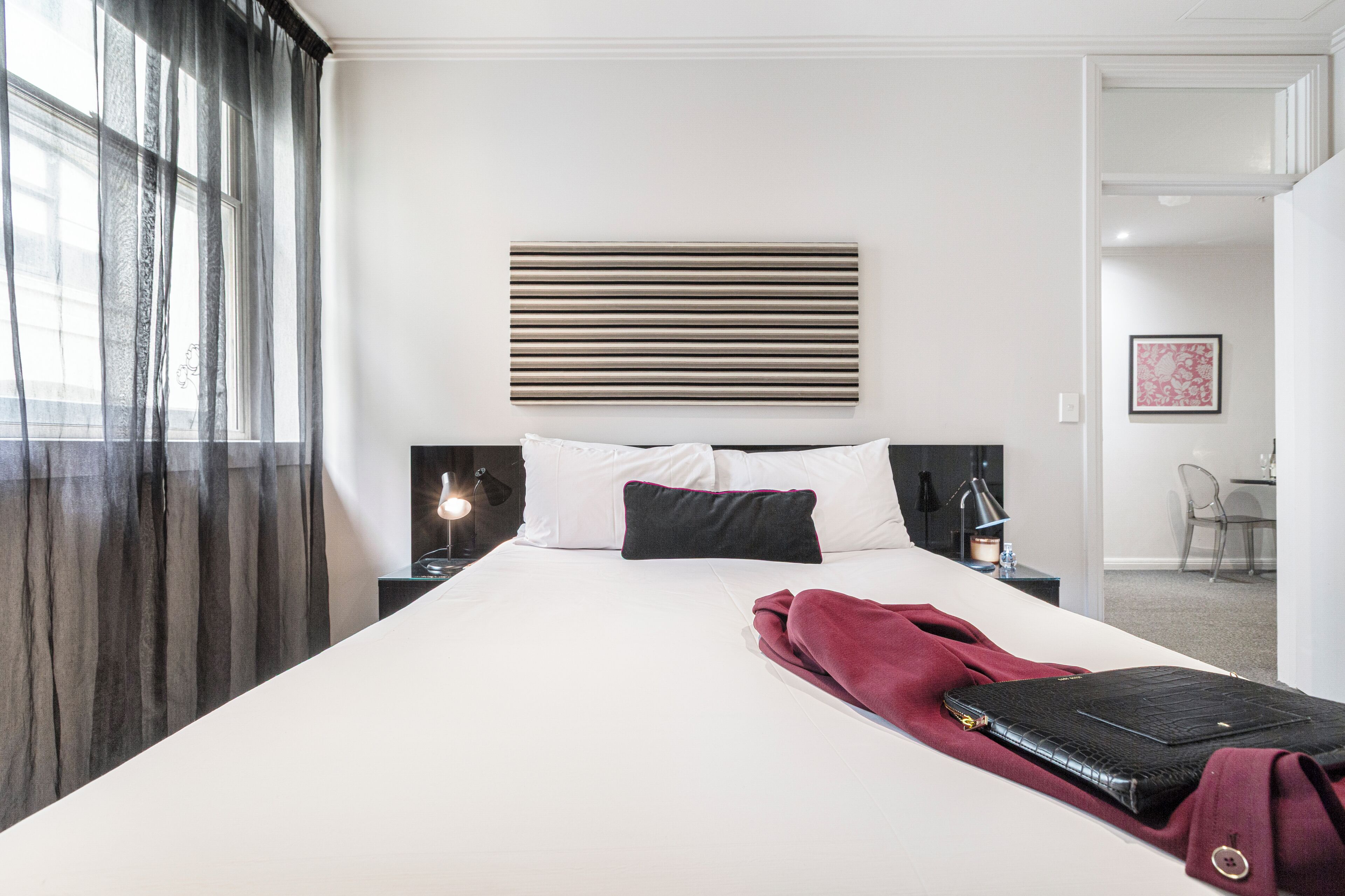Una habitación de hotel con una cama grande, un cabecero con líneas horizontales, dos lámparas de noche, una manta roja y una maleta sobre la cama.