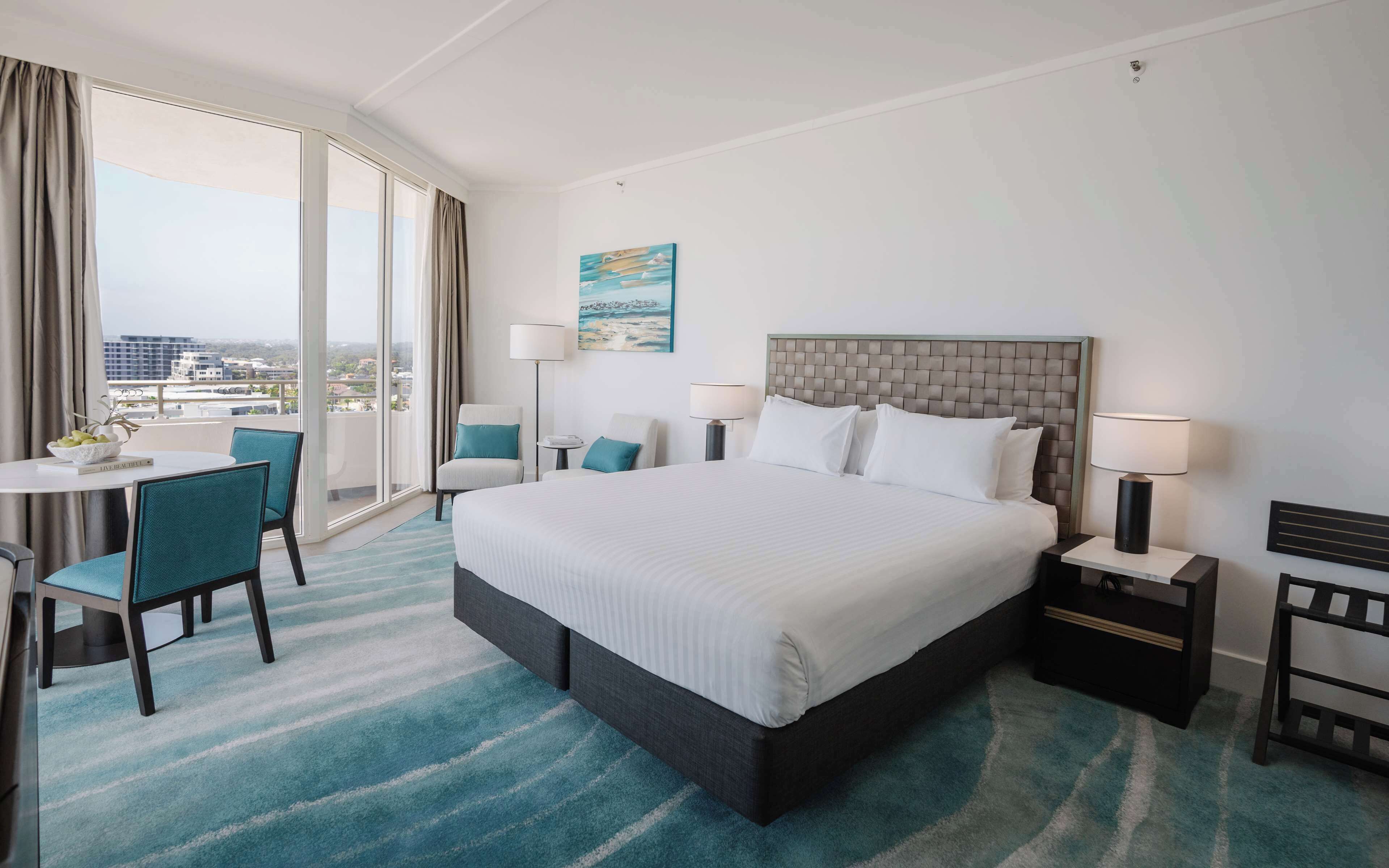 Habitación de hotel con una cama grande, un escritorio con silla, vista a la ciudad desde un balcón y un cuadro en la pared.