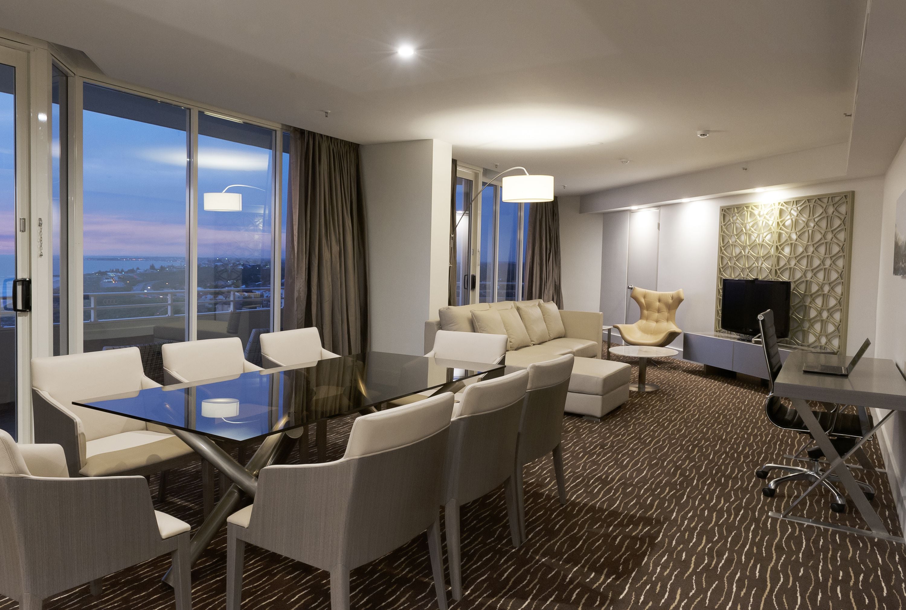 A modern hotel room with a glass dining table, white sofas, a leather armchair, a flat-screen TV, and a city view.