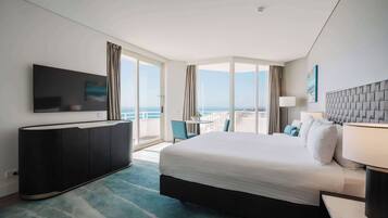 A hotel room with a large bed, a flat-screen TV mounted on the wall, a desk with a lamp, and a balcony with a view of the city.
