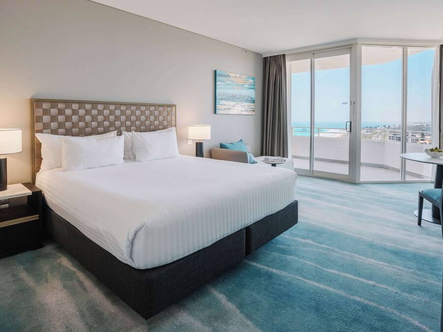 Quarto de hotel com uma cama grande, duas mesas de cabeceira com luminárias, uma mesinha com um vaso e vista para a cidade através de grandes janelas.
