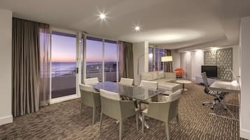 Habitación de hotel con balcón, zona de comedor, sofá, televisor y lámpara.