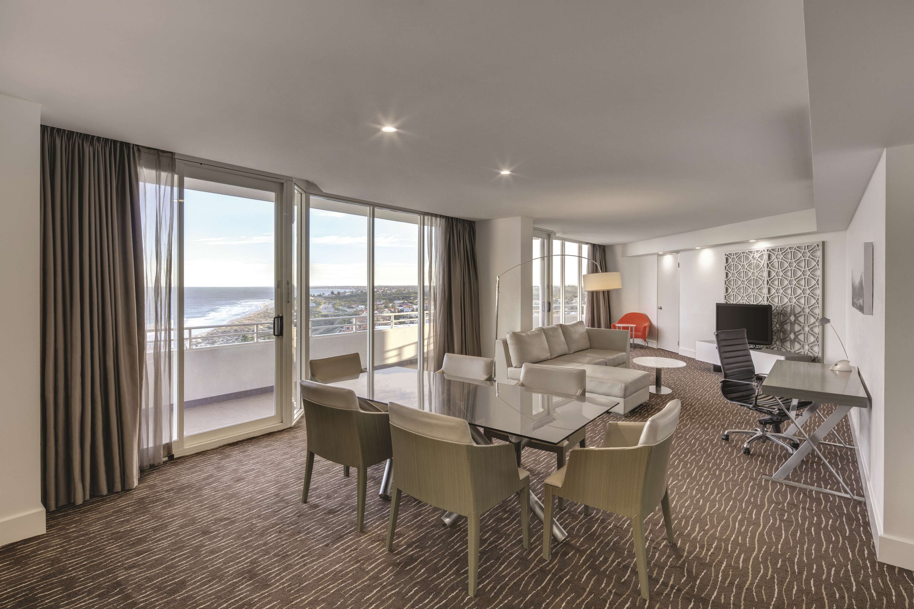 A modern hotel room with a large balcony, a dining area, a sofa, a TV, and a desk.