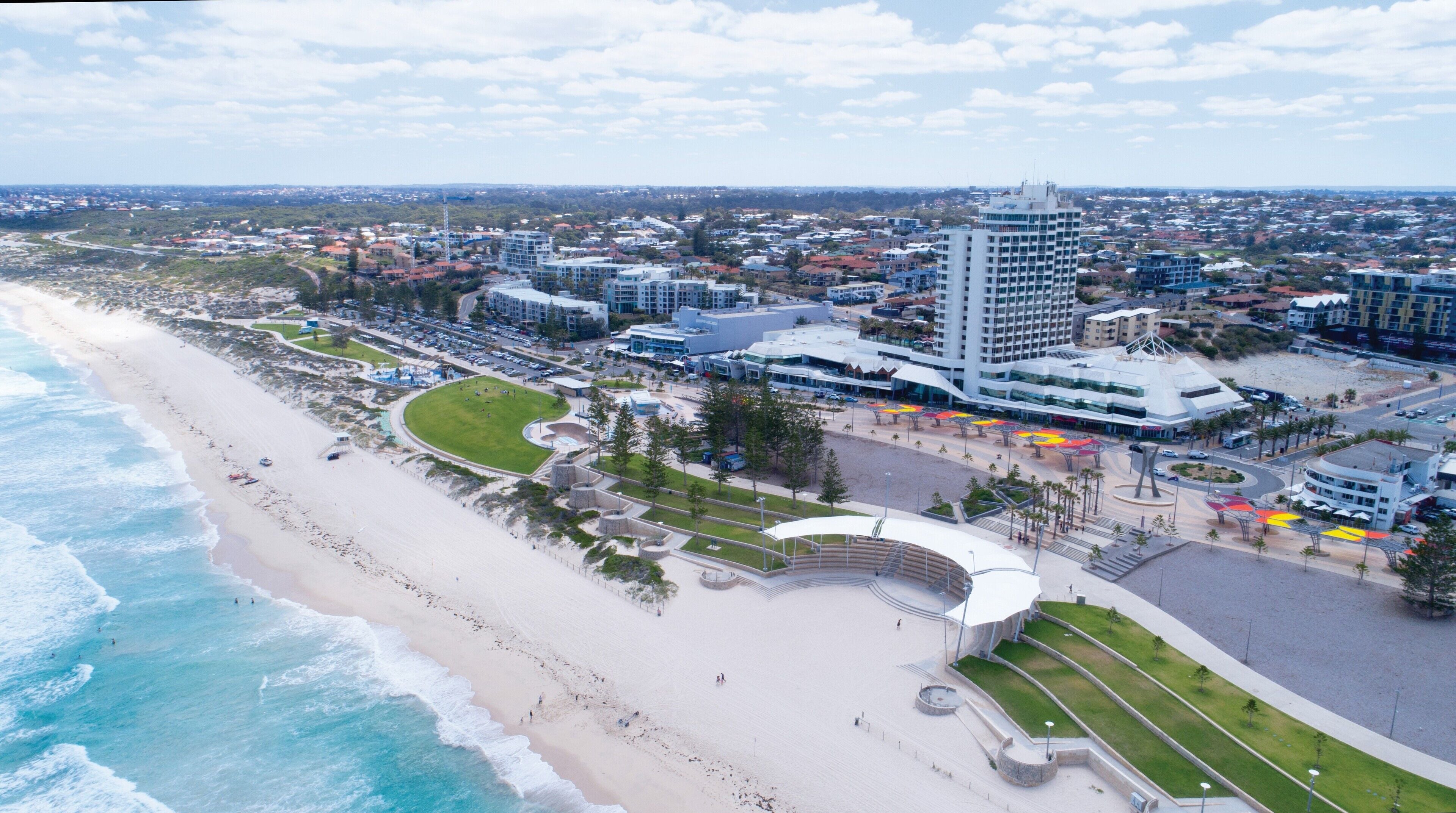 An aerial view of a coastal city with a prominent hotel featuring a modern design, a beachfront promenade, and a mix of residential and commercial buildings.