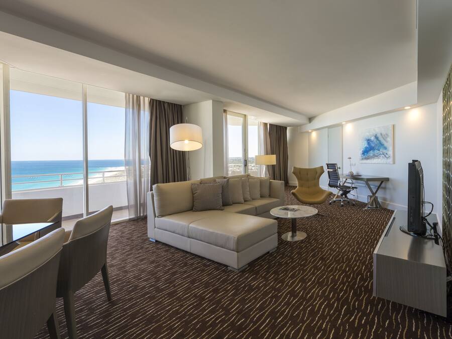 A modern hotel room with a large window offering a view of the ocean, a sofa, a small round table, and a desk with a computer.