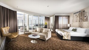 A spacious hotel room with a large bed, a sofa, a chair, and a coffee table. There is a balcony with a view of the city.