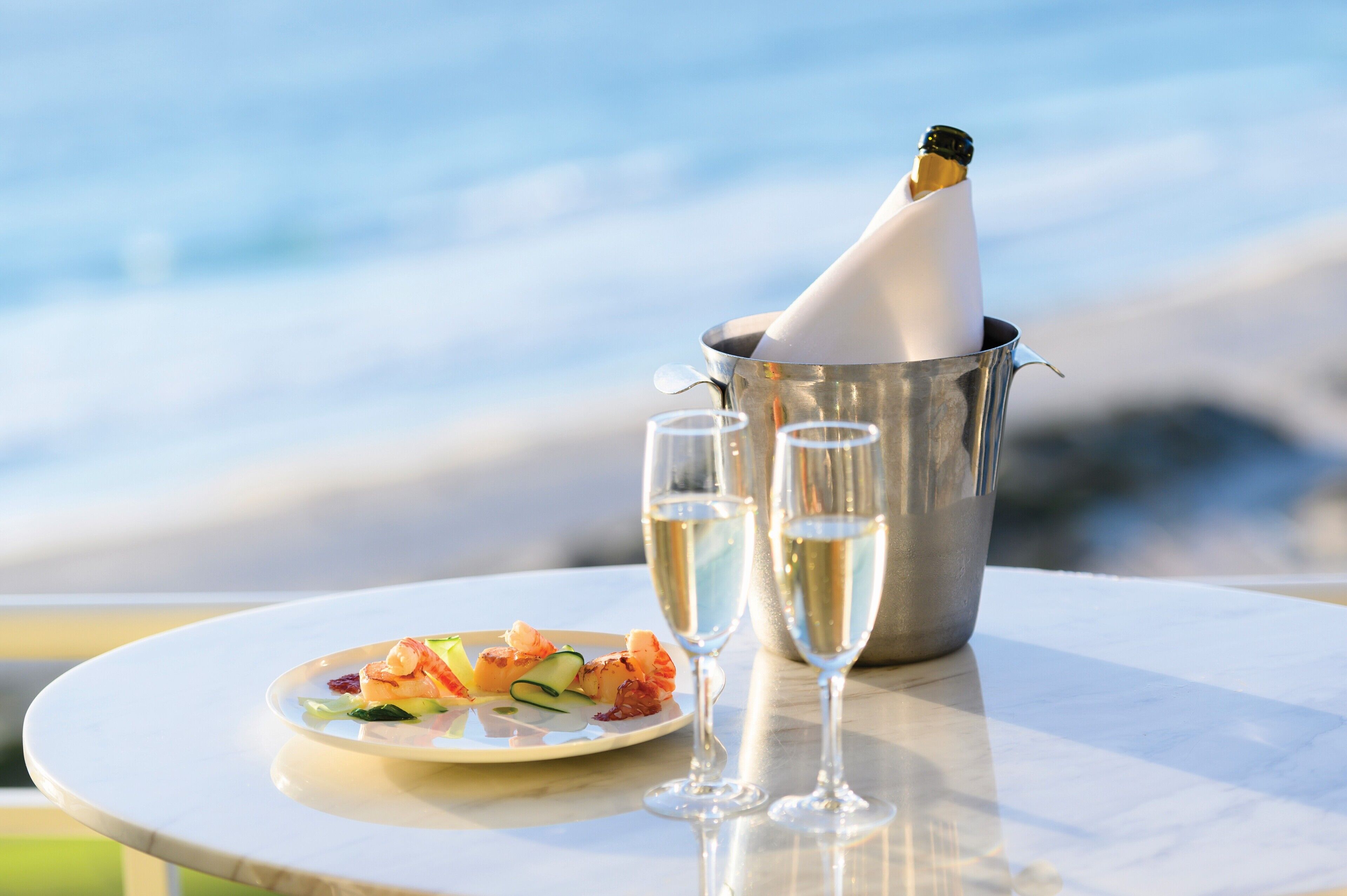 A table with a seafood dish, two champagne glasses, and a champagne bottle in a metal bucket, set against a seaside backdrop.