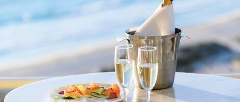 A table with a seafood dish, two champagne glasses, and a champagne bottle in a metal bucket, set against a seaside backdrop.