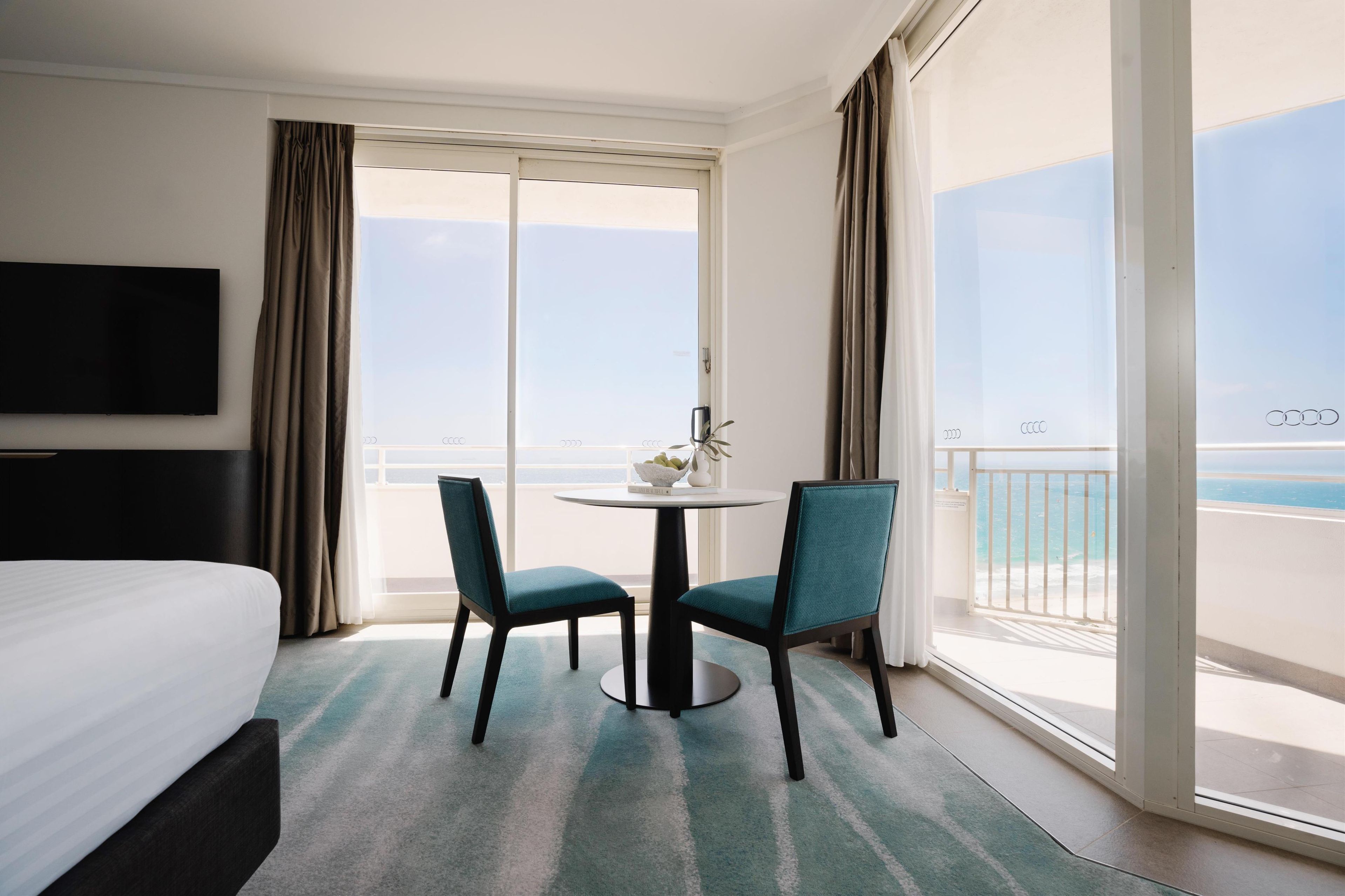A hotel room with a bed, a TV, a round table with chairs, and a balcony with a view of the sea.