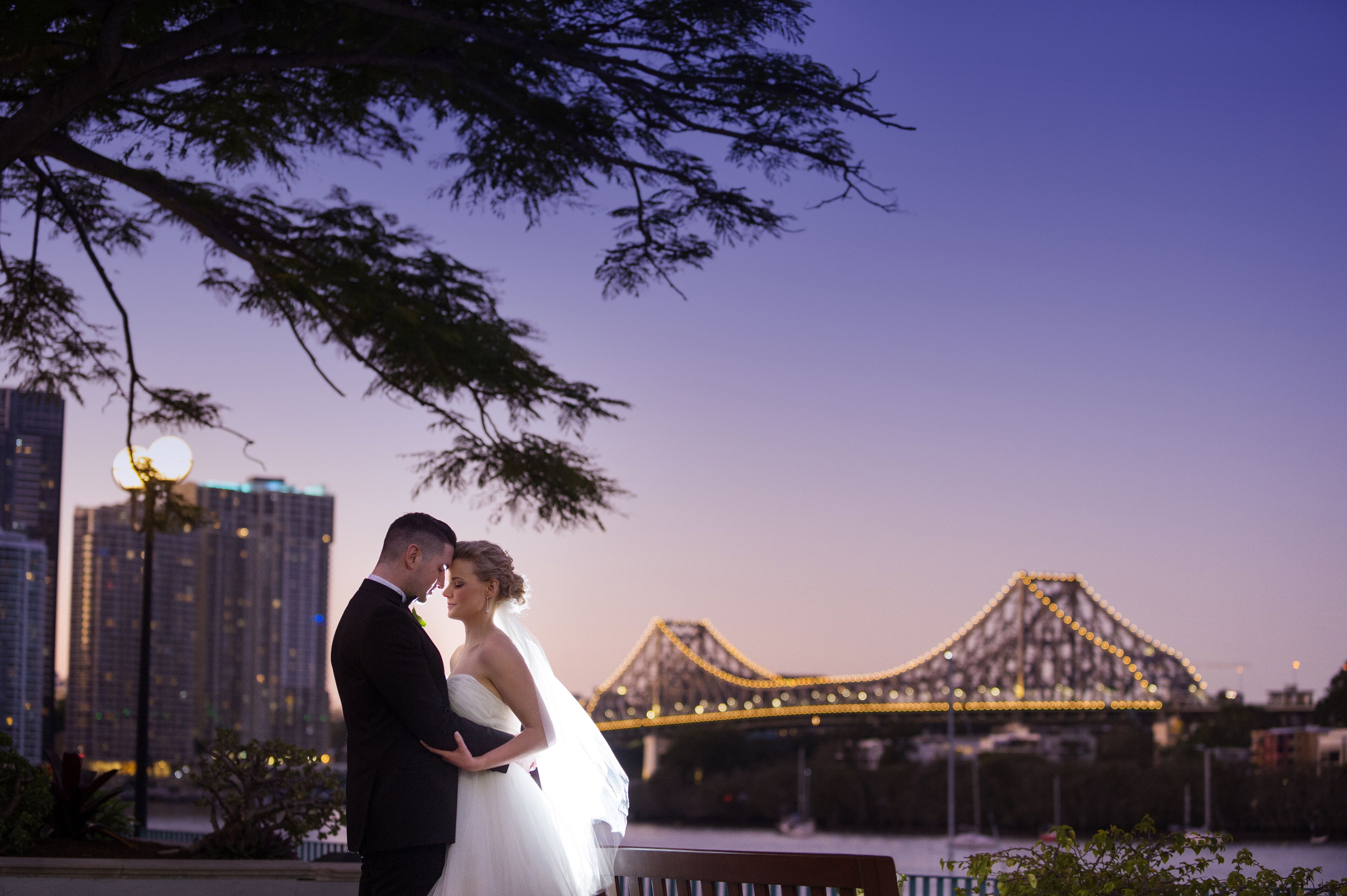 A couple embracing in front of a bridge with city buildings in the background.