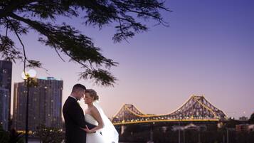 A couple embracing in front of a bridge with city buildings in the background.
