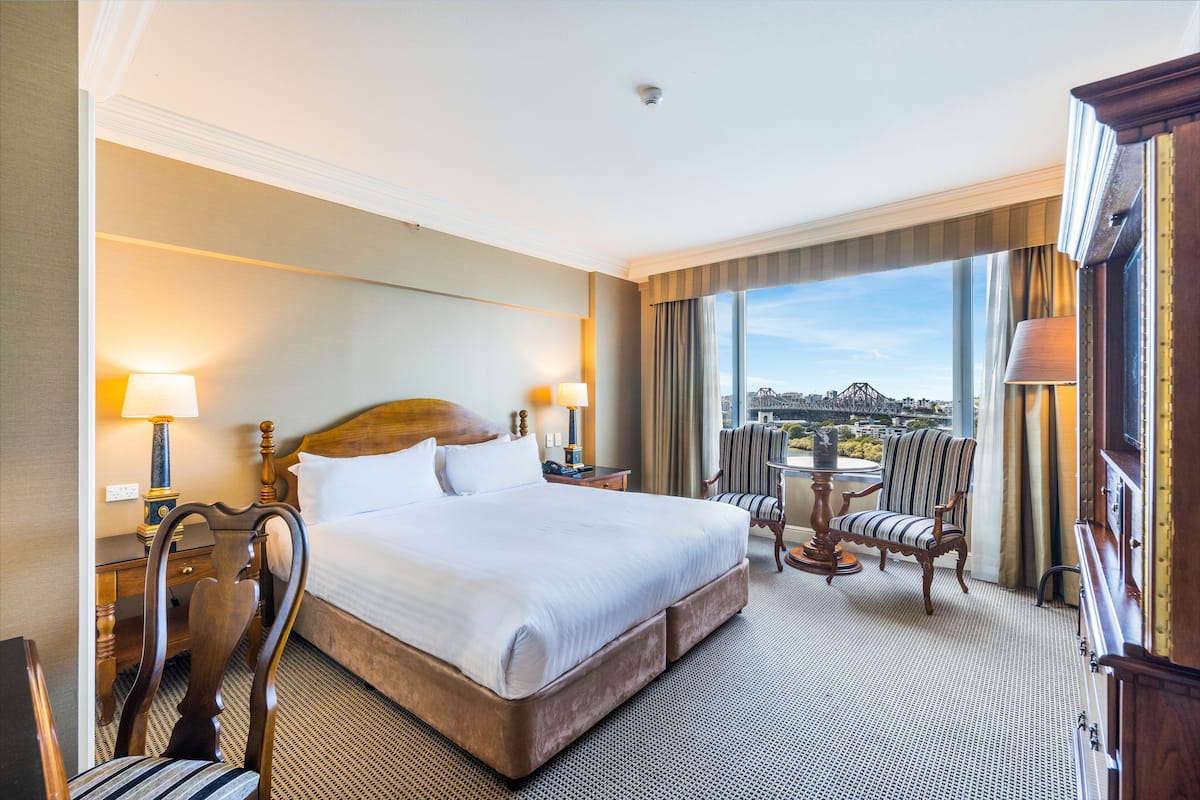 Hotelkamer met een groot bed, twee nachtkastjes met lampen, een stoel, een klein tafeltje en uitzicht op een stadsbeeld door grote ramen.