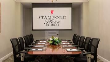 A conference room with a long table, black chairs, a floral centerpiece, and a screen displaying 'STAMFORD Plaza Brisbane' branding.