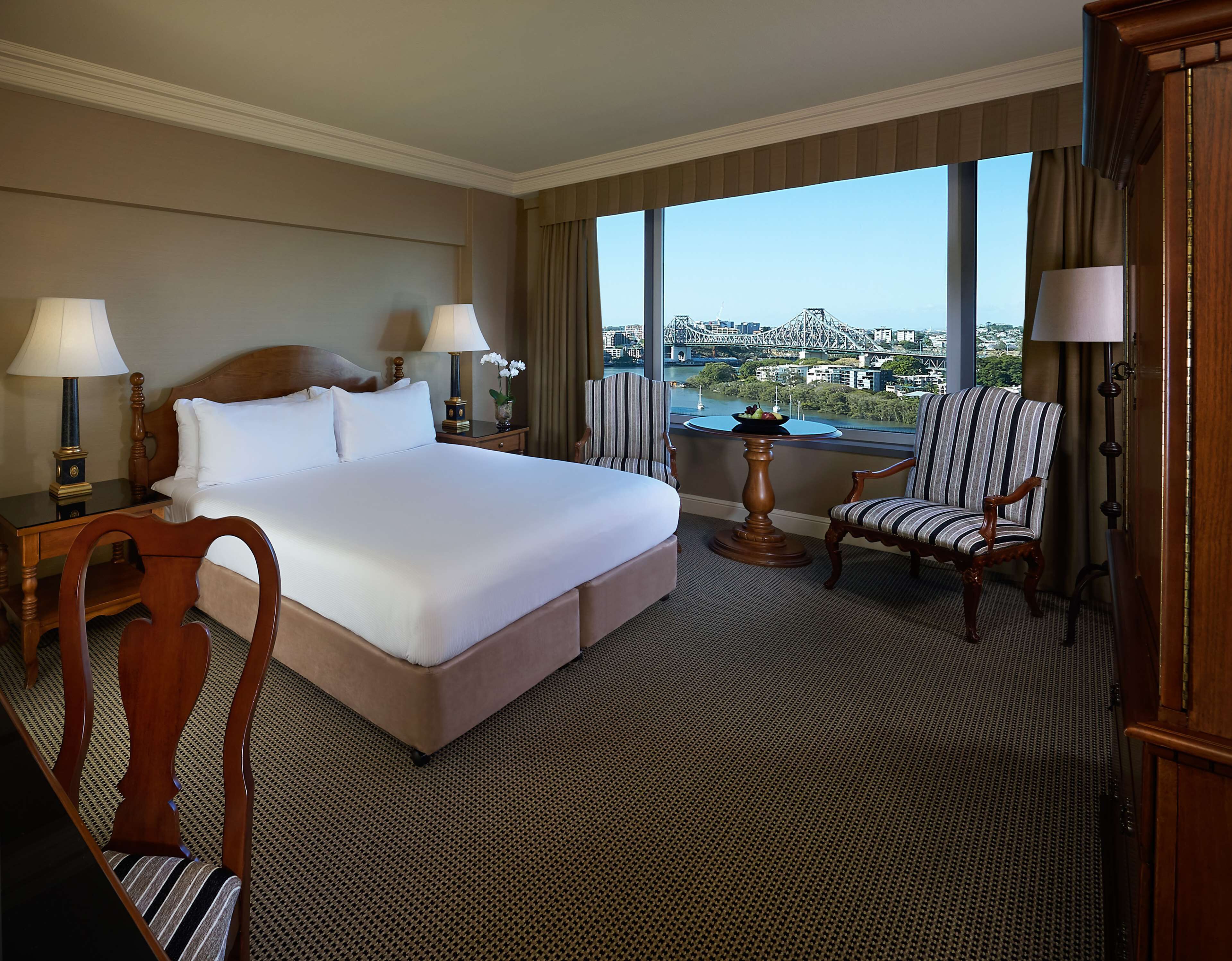 A hotel room with a large bed, two bedside tables with lamps, a chair, a round table with a fruit bowl, and a view of a cityscape.
