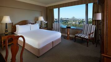 A hotel room with a large bed, two bedside tables with lamps, a chair, a round table with a fruit bowl, and a view of a cityscape.